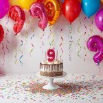 balloons and confetti in bright colors surrounding a white cake with a number 9 on top